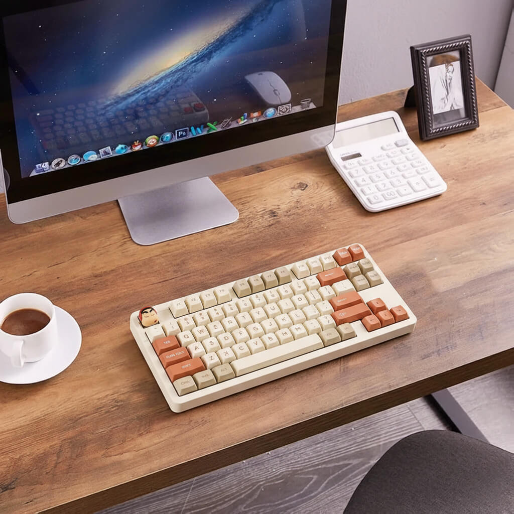 Top-down view of a desk setup featuring a Crayon Shin-chan artisan keycap on the ESC key of a mechanical keyboard, with a Mac computer and coffee cup nearby
